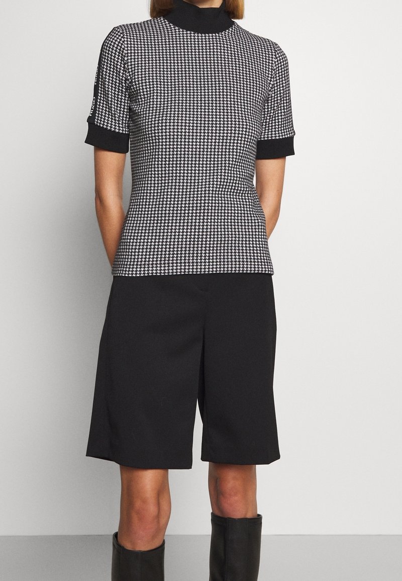 Czarny i biały sweter w wzór houndstooth z krótkim rękawem i wysokim dekoltem, w połączeniu z czarnymi szortami o szerokich nogawkach oraz czarnymi botkami sięgającymi za kolano.
