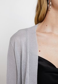 Silver lättviktscardigan med ett strukturerat stickmönster, öppen framsida och mjukt, skimrande tyg som hänger över en svart satintopp.