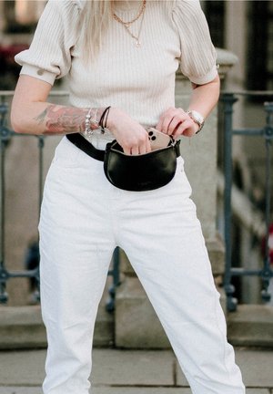 Marsupio in pelle nera con chiusura a zip, indossato in vita, posizionato davanti a una persona che indossa jeans bianchi e una maglia beige a coste.