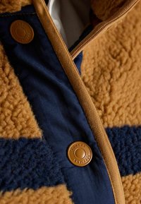 Brūnas un tumši zilas jakas ar teksturētu vilnas ķermeni, gludas auduma apdari un apaļu bronzas pogu ar iegriezto logo.