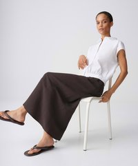 Woman wearing a white sleeveless top, long dark skirt, and flip-flops sits sideways on a white chair against a plain background.