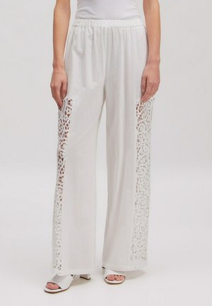 Weiße weit geschnittene Hosen mit floralen Spitzeneinsätzen an den Außenseiten, getragen mit weißen offenen Sandalen und einem weißen ärmellosen Oberteil.
