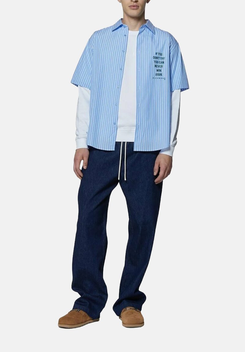 Camicia a maniche corte a righe blu sopra una camicia a maniche lunghe bianca, abbinata a pantaloni di jeans scuri e scarpe slip-on marroni. Testo sulla camicia che legge.