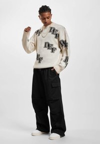 Cremiger, oversized Sweater mit schwarzen "DEF" Logos, kombiniert mit lockeren schwarzen Cargo-Hosen und weißen Sneakers. Sanfte Textur und lässige Passform.