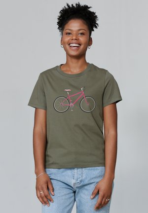 T-shirt vert olive avec un graphisme de bicyclette rose. Manches courtes, encolure ronde, fabriqué en tissu doux, porté avec un jean bleu clair.