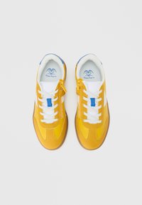 Para żółtych tenisówek z białymi sznurowadłami, niebieskimi akcentami, bocznymi suwakami i sercowatym logo wewnątrz, widocznych z góry na białym tle.