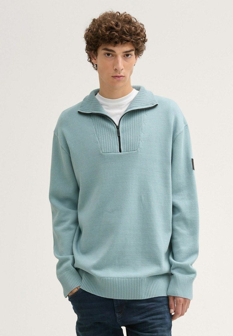 Hellblauer Strickpullover mit einem Reißverschlusskragen und gerippten Bündchen. Besitzt ein strukturiertes Paneel am Hals und einen lässigen Schnitt.