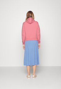 Růžový zkrácený mikina s kapucí, spárovaná s modrou midi sukní s žebrováním a teniskami v neutrálních tónech, viděná ze zadní strany.