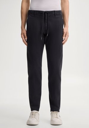 Homme portant un pantalon noir avec cordon et des baskets blanches, debout devant un fond clair uni.