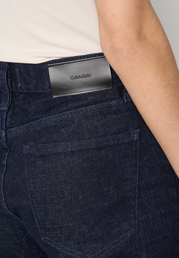 Ciemnoniebieskie dżinsy o gładkiej fakturze, z czarną skórzaną naszywką w pasie z wytłoczonym napisem "Calvin Klein". Widoczna tylna kieszeń.