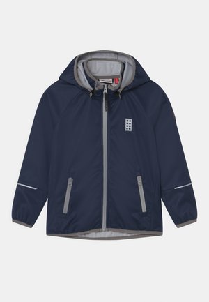 Veste coupe-vent bleu marine avec des accents gris, devant zippé et capuche. Comprend deux poches latérales et des détails réfléchissants sur les manches.