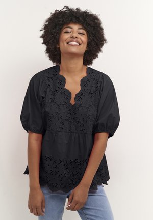Schwarze Bluse mit floralem Lochstickerei, Puffärmeln und einem gewellten Ausschnitt. Weicher Stoff, geschichtetes Design; kombiniert mit hellen Jeans.