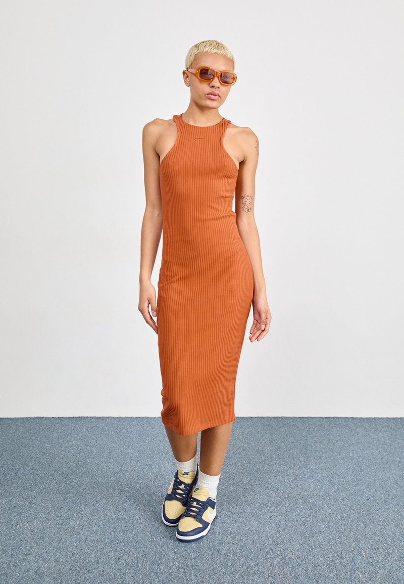 Vestido midi naranja ajustado con ribetes y cortes en los hombros. Combinado con zapatillas amarillas y marinas y calcetines blancos, sobre un fondo de alfombra azul.
