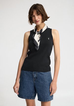 Femme portant un haut noir sans manches à côtes, un short en jean bleu et une écharpe noire et blanche nouée autour du cou.