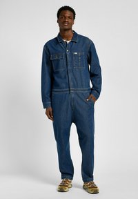 Combinaison en denim bleu foncé, avec fermeture éclair à l'avant, deux poches poitrine, manches longues et coupe décontractée. Portée avec des chaussures de couleur claire.