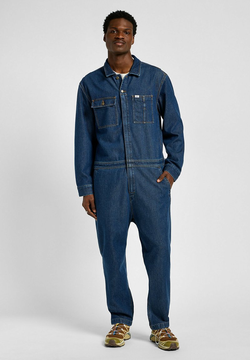 Combinaison en denim bleu foncé, avec fermeture éclair à l'avant, deux poches poitrine, manches longues et coupe décontractée. Portée avec des chaussures de couleur claire.