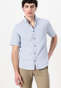 Camicia con maniche corte e bottoni di colore blu chiaro, con un motivo a texture, bottoni neri e una tasca sul petto anteriore. Tessuto di cotone liscio.