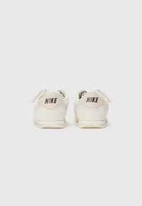 Et par hvite Nike-sneakers med beige accenter vist fra baksiden, som viser Nike-logoen på hæltabbene, mot en enkel hvit bakgrunn.