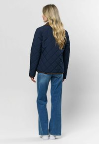 Tamsiai mėlynas siuvinėtas paltas su rudomis apykaklėmis, ilgomis rankovėmis ir tiesiu siluetu, derinamas su šviesiai mėlynomis platėjančiomis džinsomis ir baltais sportbačiais.