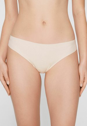 Frau trägt nahtlose, hellbeige Bikini-Unterwäsche, steht mit entspannten Armen an den Seiten vor einem schlichten Hintergrund.