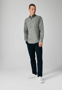 Graues Langarm-Hemd mit Knöpfen und Brusttasche, kombiniert mit navyfarbener Hose und weißen Sneakers. Es hat einen kragenartigen Hals und eine subtile Textur.