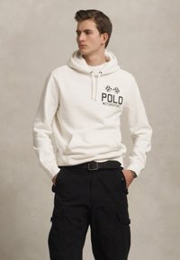 Balta kapuce ar priekšējo kabatu, kurā ir melna "POLO MOTORSPORT" teksta un šaha karoga dizains. Apvienota ar melnajiem darba biksēm.