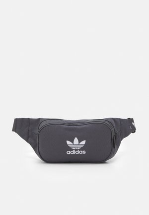 Zwarte Adidas heuptas met een voorvak met rits en wit trefoil-logo in het midden van het tasje tegen een effen achtergrond.
