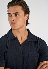 Donkerblauw poloshirt met een gestructureerde stof, korte mouwen, een puntige kraag en een borstzakje. Model heeft een gesloten, neutrale uitdrukking.