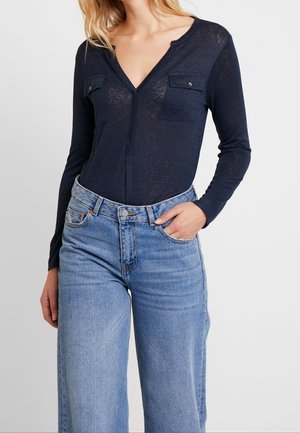 Woman wearing a navy blue V-neck long-sleeve top with chest pockets and light blue high-waisted wide-leg jeans, hand in pocket.