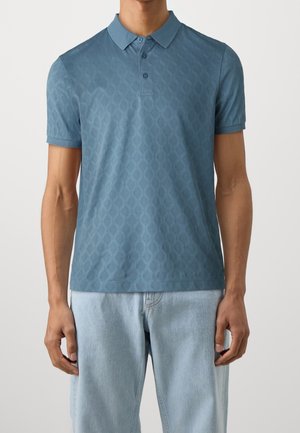 Man draagt een blauw poloshirt met korte mouwen en patroon en lichtblauwe spijkerbroek, staand met ontspannen armen langs zijn zij.
