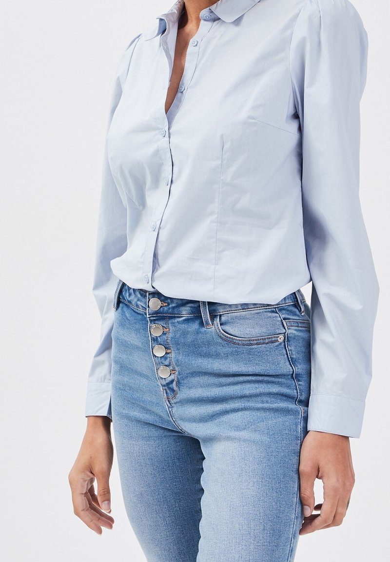 Chemise bleu clair à boutons avec col et manches bouffantes, associée à un jean taille haute bleu avec des accents de boutons argentés.