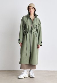 Long, olive green waterproof coat with a high collar, front zipper, belted waist, and black accents. Worn over a beige top and brown skirt.