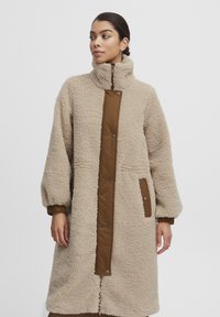 Fleece lång kappa i ljusbeige med hög krage, brun knäppt frampanel, ribbade ärmslut och sidofickor. Mjuk, texturerad yta.