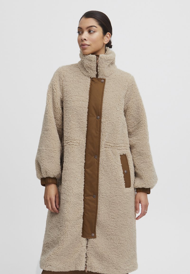 Fleece lång kappa i ljusbeige med hög krage, brun knäppt frampanel, ribbade ärmslut och sidofickor. Mjuk, texturerad yta.