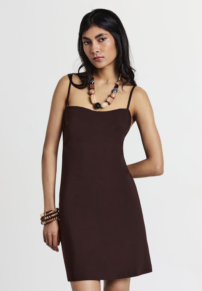 Braunes, tailliertes Kleid mit dünnen Trägern und geradem Ausschnitt. Das Model hat eine dicke Halskette aus mehrfarbigen Perlen und Holzbändern accessorisiert.