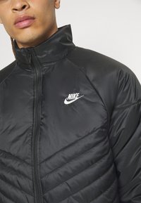 Giacca puffer nera Nike con colletto alto, dotata di chiusura con zip e logo ricamato bianco sul petto, che presenta una texture trapuntata.