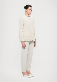 Blazer beige con texture, collo rotondo, cinque bottoni e due tasche frontali, abbinato a pantaloni dritti crema chiaro e mule a punta.