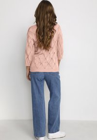 Helles rosa gestricktes Sweatshirt mit einem Rautenmuster, 3/4-Ärmeln, kombiniert mit blauen weit geschnittenen Jeans und weißen Sneakers.