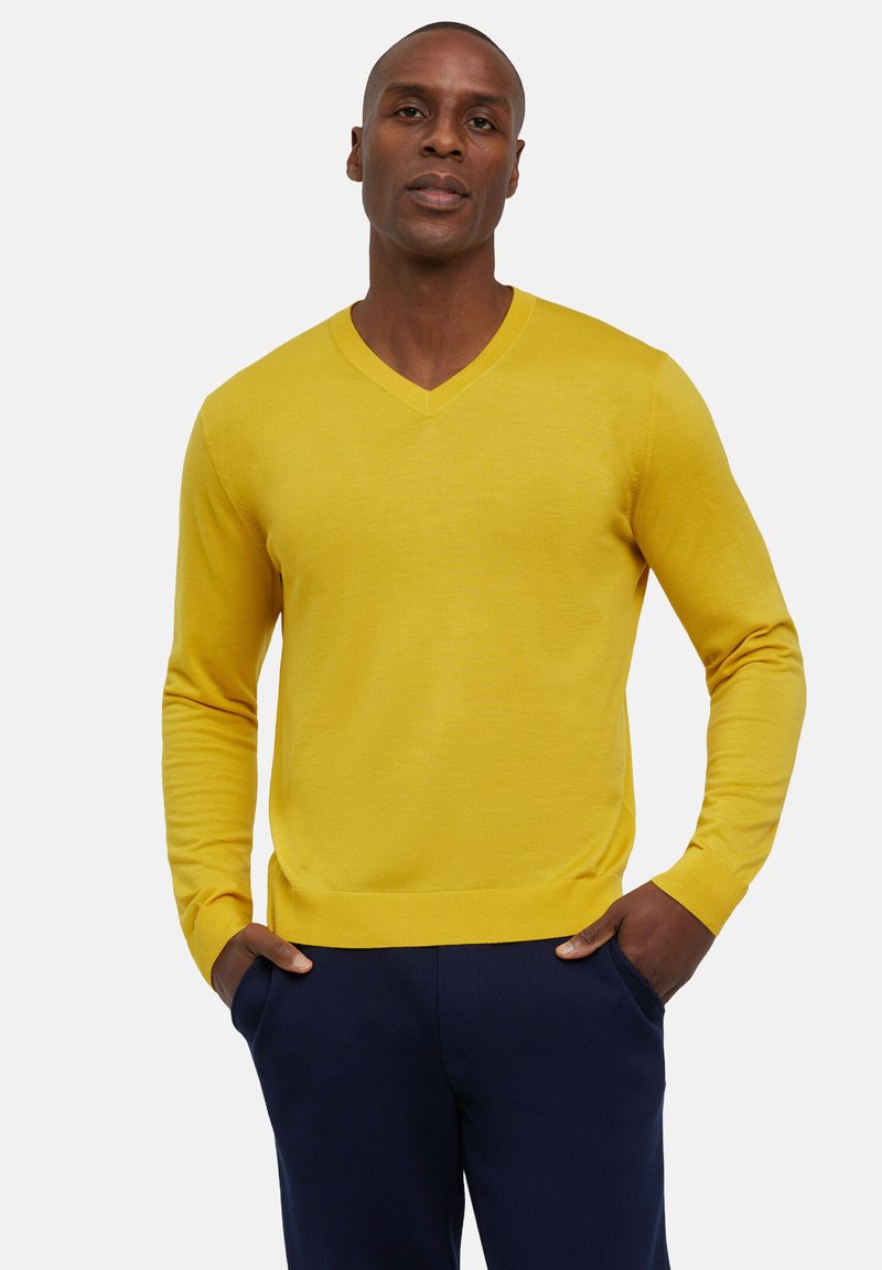 Gelbes V-Ausschnitt-Pullover mit langen Ärmeln, aus glattem Stoff gefertigt, kombiniert mit dunklen Marinehosen, die ein einfaches, klassisches Design betonen.