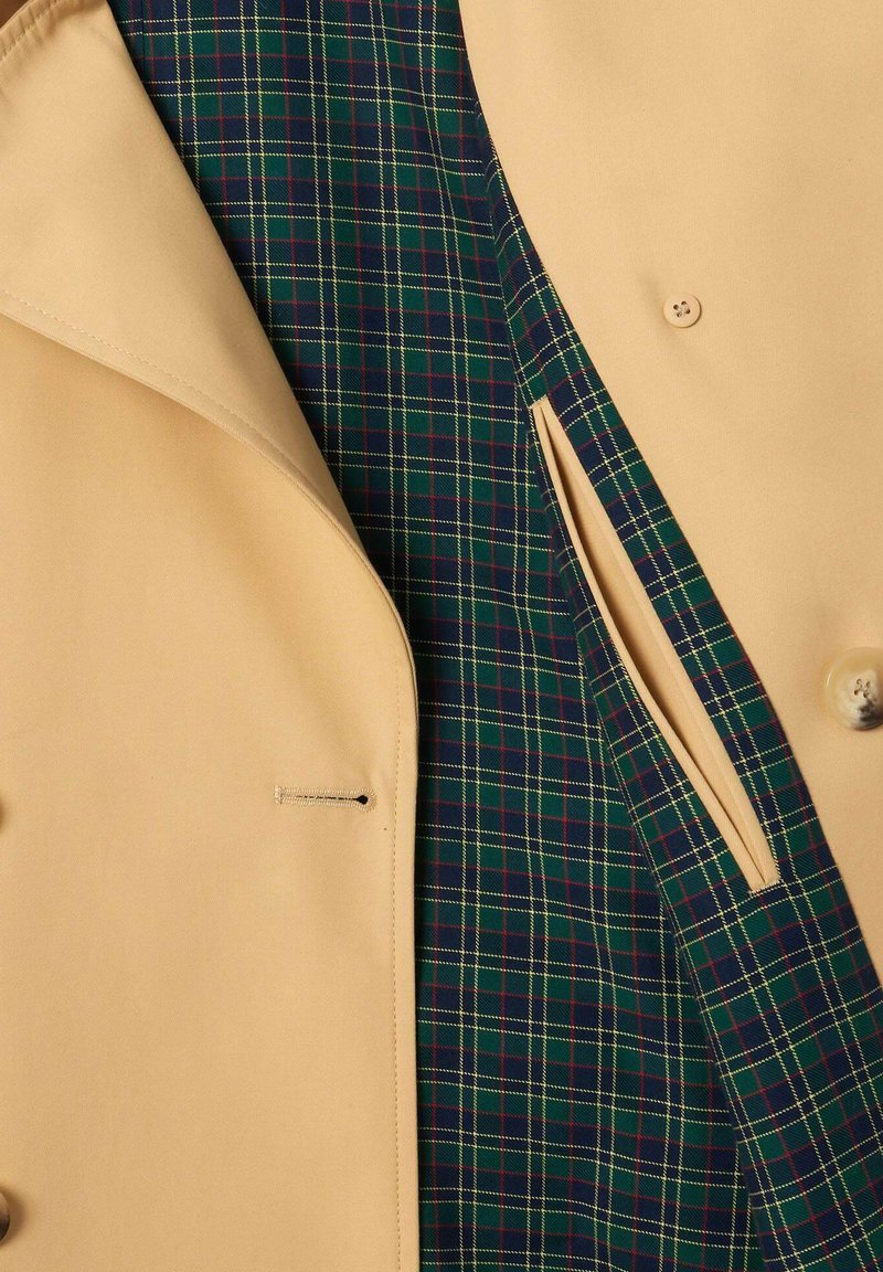 Manteau beige avec une doublure intérieure à carreaux dans des tons de vert, bleu marine et rouge. Texture lisse avec une fermeture à un bouton et des poches.