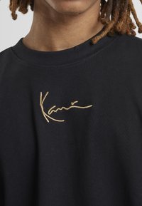 Acercamiento de una persona que lleva una camiseta negra con el logo dorado bordado "Karni" en el pecho y rastas marrones visibles.