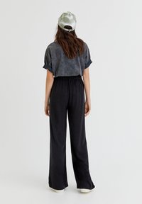 Kobieta z długimi brązowymi włosami, ubrana w szaro-wyblakły t-shirt, czarne szerokie spodnie, białe buty i jasnoszary czapkę bejsbolówkę, odwrócona plecami.