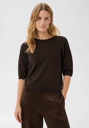 Femme blonde portant un haut en maille noir à manches courtes avec des bords festonnés et un pantalon marron foncé, debout avec une main dans la poche.