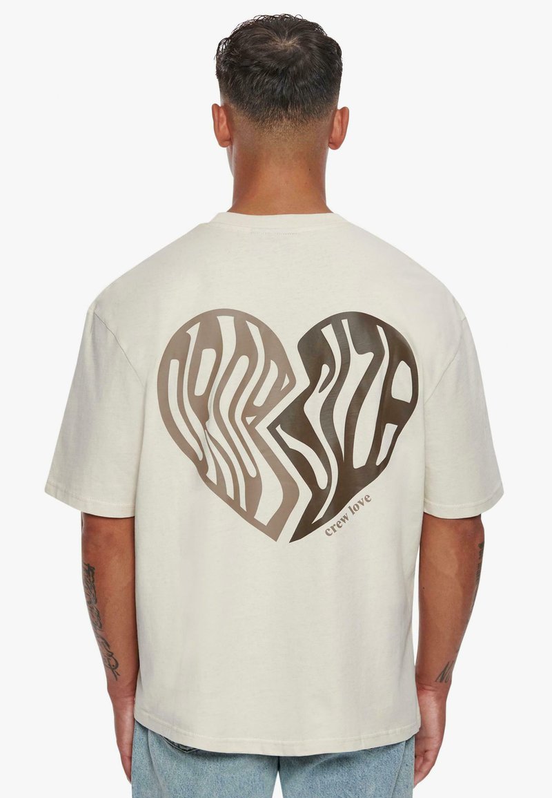 Kurzärmliges cremefarbenes T-Shirt mit einem großen Herz-Graphic, das mit wellenförmigen braunen Linien verziert ist, darunter der Schriftzug "crew love". Hergestellt aus weichem Baumwollmaterial.