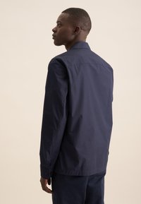 Maglietta a maniche lunghe blu navy realizzata in tessuto liscio, con colletto a punta, polsini abbottonati e vestibilità sartoriale, mostrata da un'angolazione laterale.