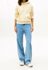 Kermanhuppari, jossa on etutasku, yhdistettynä vaaleansinisiin leveisiin farkkuihin. Kengät ovat vaaleanharmaat teksturoidulla pohjalla.