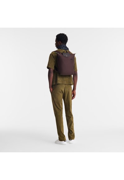 Brown backpack with a minimalist design, featuring a rounded top, top handle, and adjustable shoulder straps, worn over an olive green outfit.