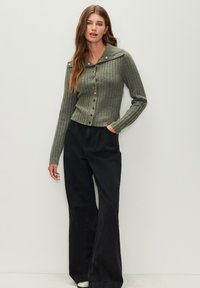 Grüner, gerippter Cardigan mit Kragen, Knopfverschluss und dekorativen goldfarbenen Knöpfen, kombiniert mit schwarzen, weit geschnittenen Hosen.