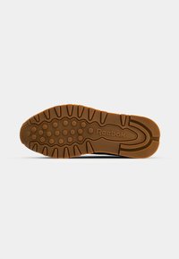 Svart skosåle med en teksturert gummioverflate, preget av sirkulære og lineære mønstre. "Reebok"-logoen er preget i midten. Brun traksjonsyttersåle.