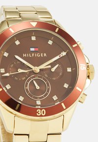 Reloj de muñeca dorado con esfera marrón, bisel rojo y marcadores de diamante. Presenta subesferas para el día y la hora en formato de 24 horas. Banda de acero inoxidable.
